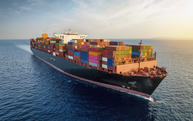 Aerial view of a large, loaded container cargo ship traveling over open ocean during sunset time